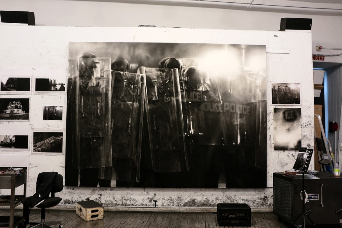 Black and white photo of police in riot gear displayed on a studio wall surrounded by smaller images and art supplies.