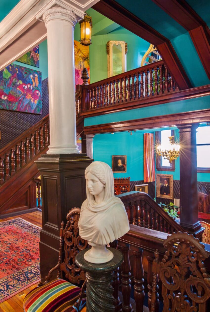 Bust on pedestal in colorful historic interior with ornate wood staircase, vibrant walls, and eclectic decor.