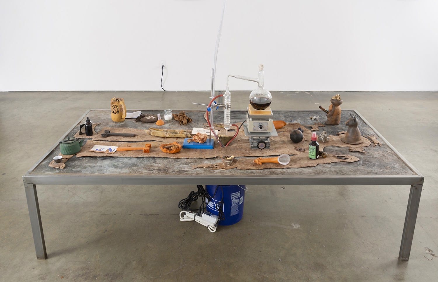 Art installation on a metal table featuring various objects, scientific equipment, and tools arranged on a textured surface.