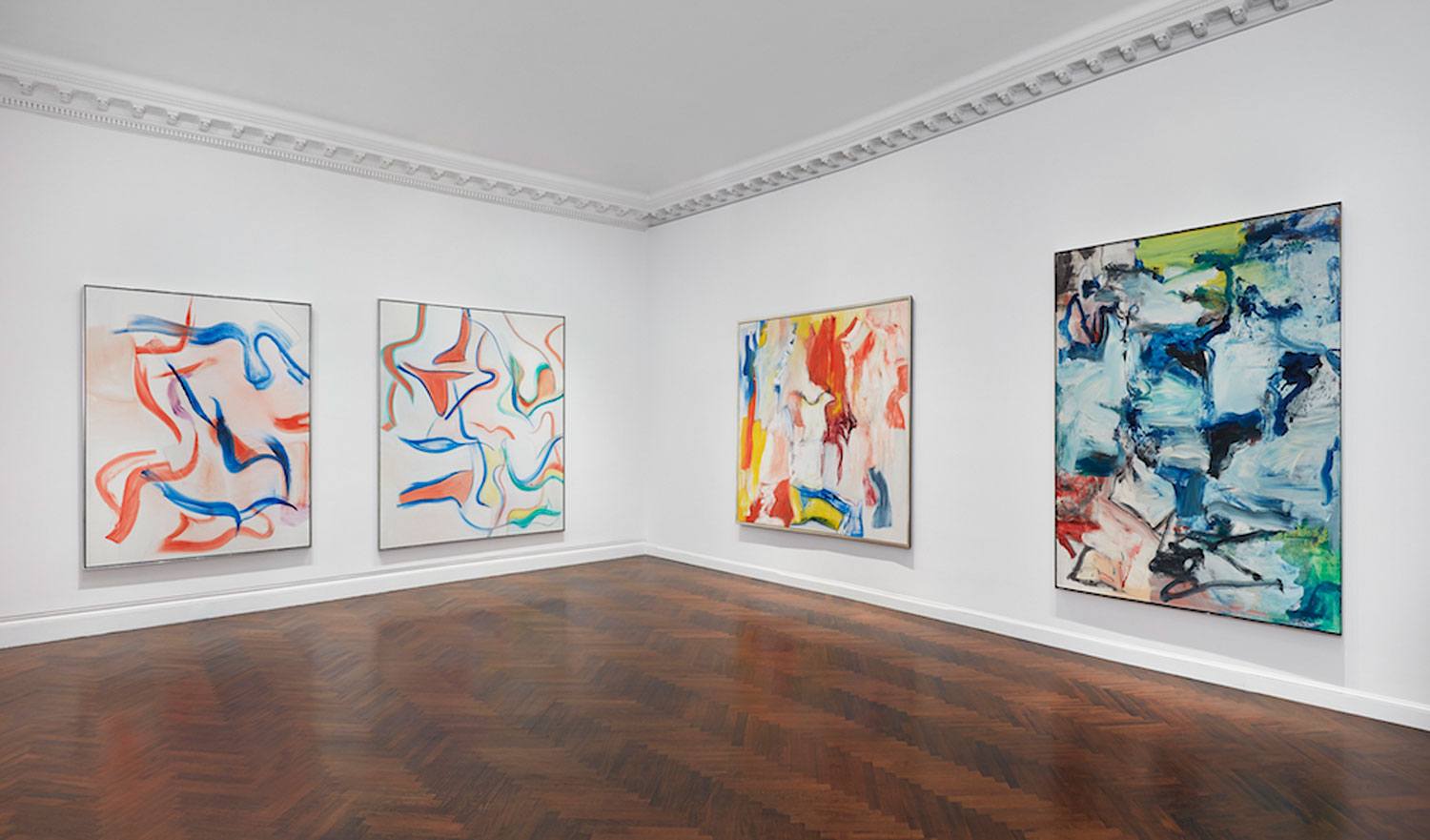Contemporary art gallery room displaying abstract paintings on white walls with wooden parquet flooring.