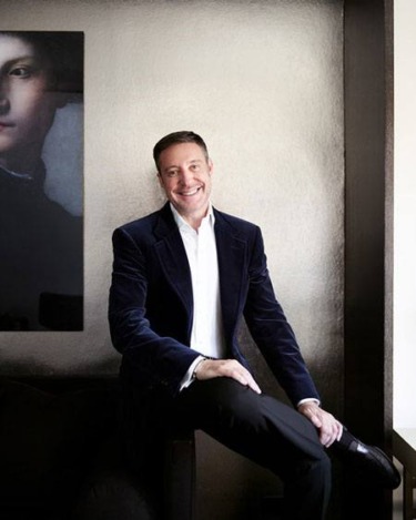 Person in a dark suit sitting on a chair with a painting in the background, smiling and looking at the camera.