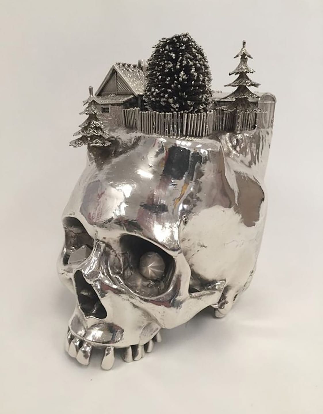 Silver skull sculpture with miniature house, trees, and fence on top, set against a plain white background.