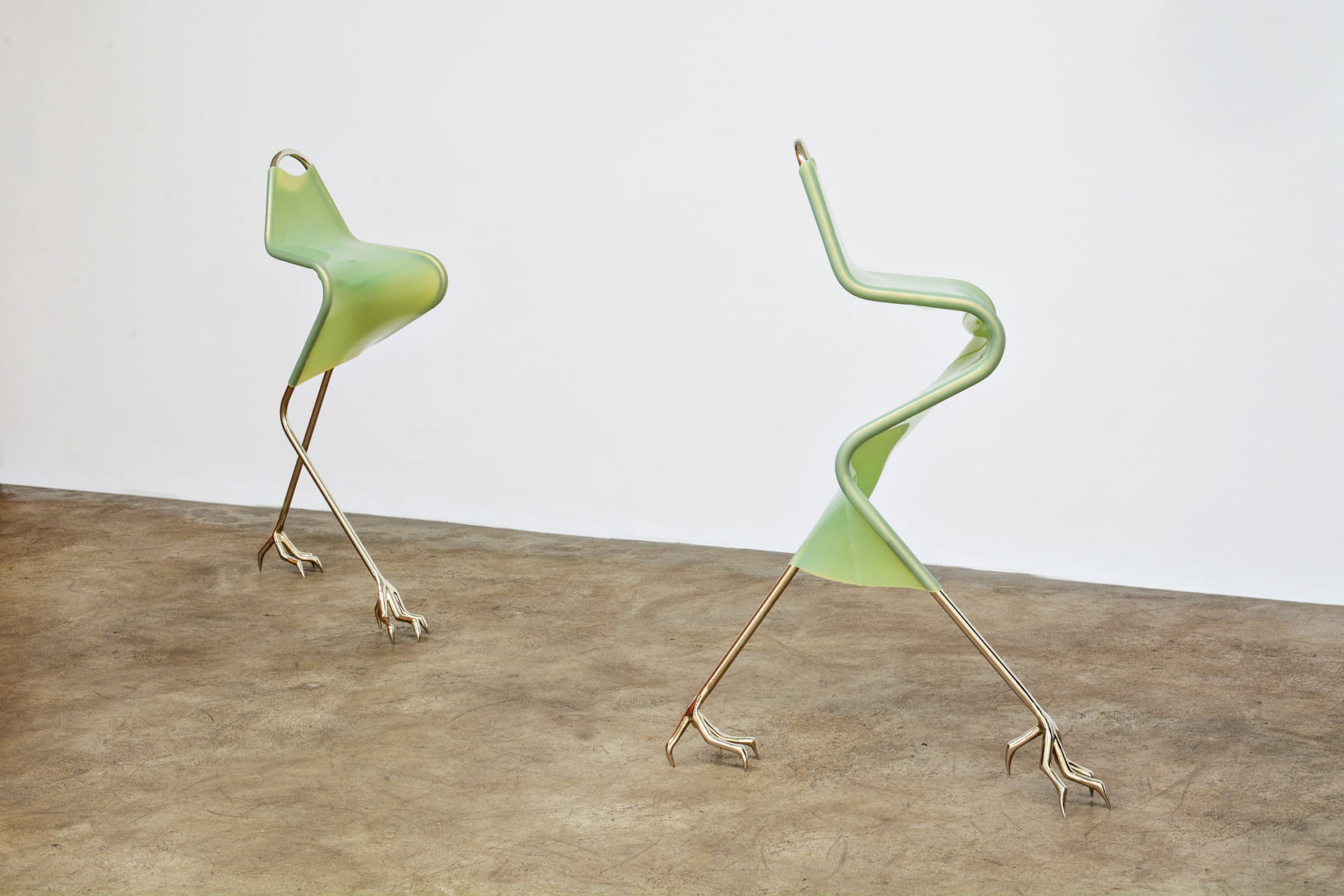Two abstract, green chairs with bird-like legs on a concrete floor, resembling modern art sculptures.
