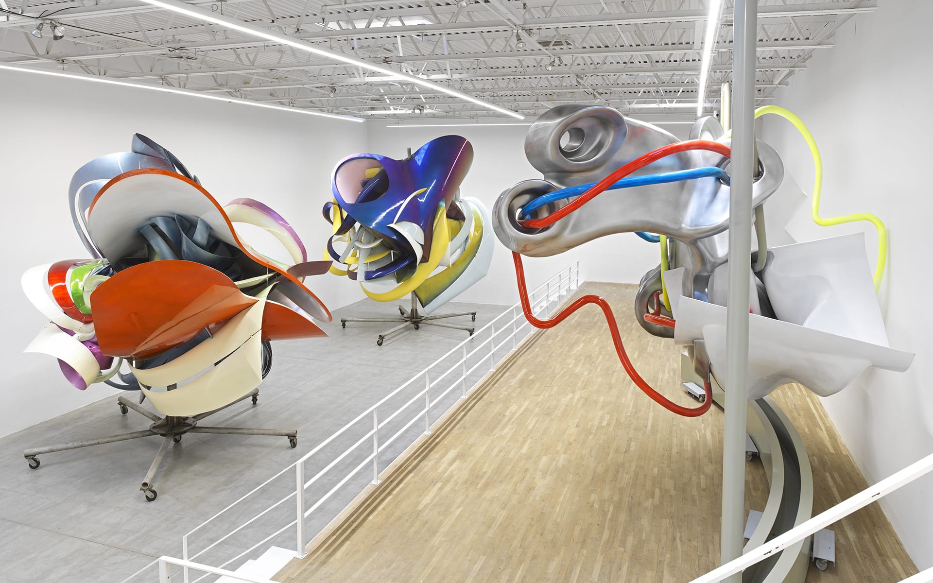 Large abstract metal sculptures in a spacious white gallery with wooden floors and a high ceiling, featuring vivid colors.