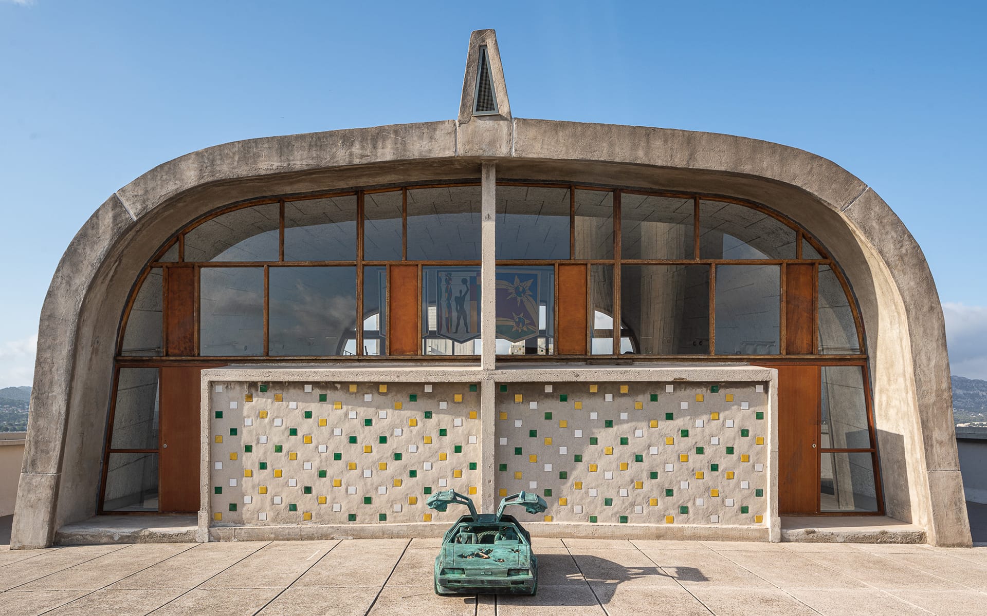 Modernist church facade with colorful tile wall and a turquoise sculpture in front under a clear blue sky.