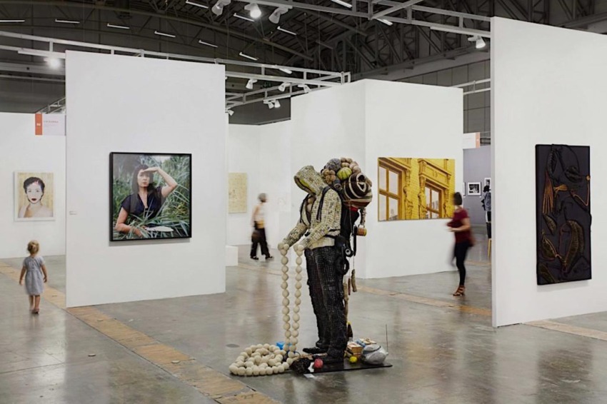 Art exhibition with sculptures and framed paintings displayed in a spacious gallery, visitors walking and observing artworks.