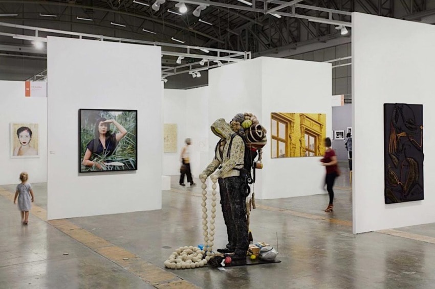 Art exhibition with sculptures and framed paintings displayed in a spacious gallery, visitors walking and observing artworks.