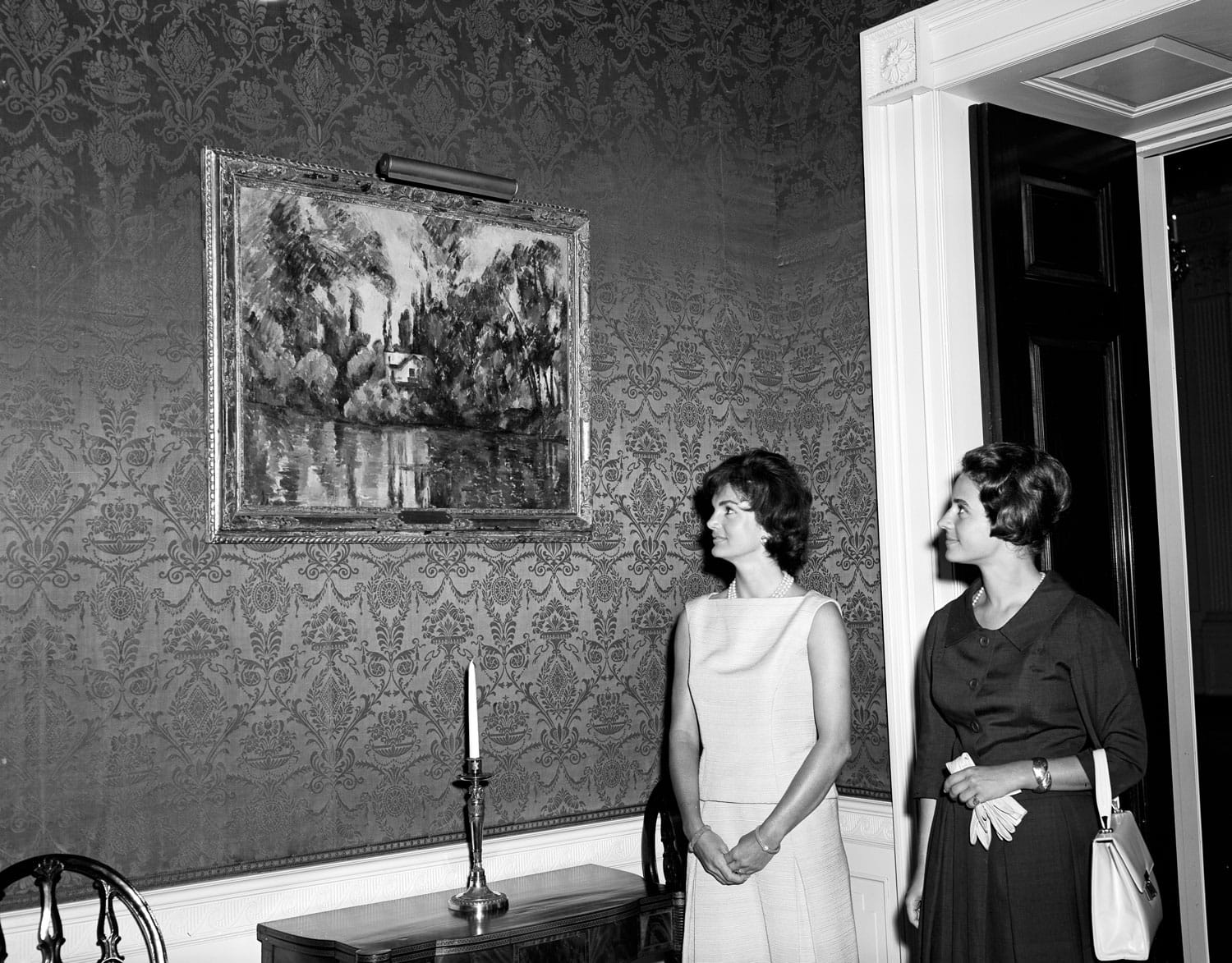 Two women in a room with patterned wallpaper looking at a framed painting hanging on the wall above a table with a candle.