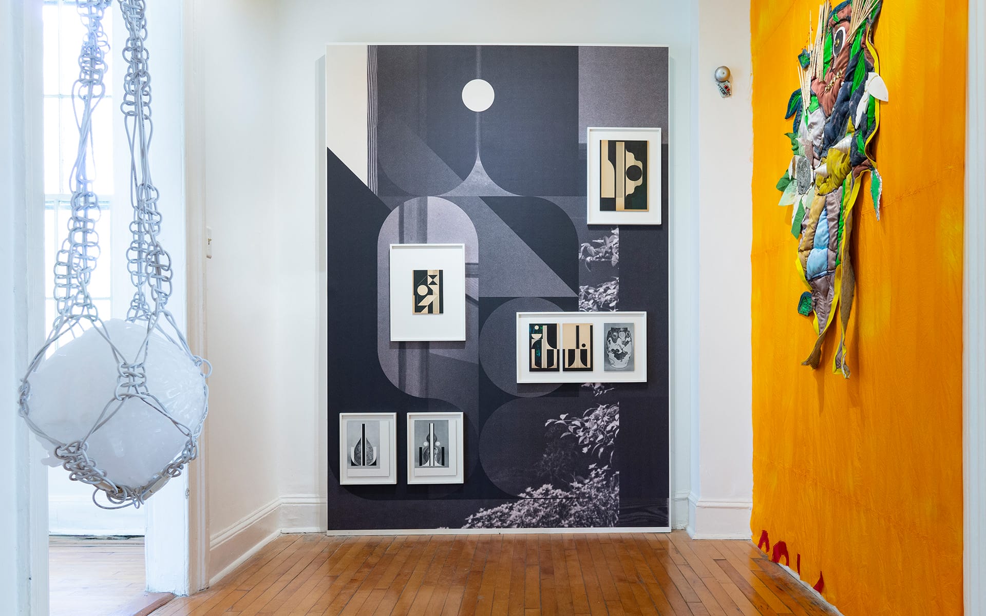 Art gallery hallway with framed abstract art on a monochrome wall and colorful textile art on the adjacent bright orange wall.