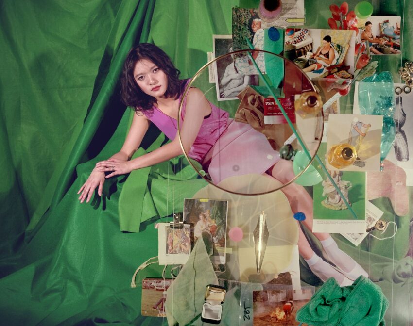 A woman in a pink dress sits on green fabric surrounded by various vintage photographs and objects.