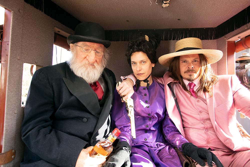 Three people in vintage clothing sitting inside a carriage, one holding a bottle, with expressions of contemplation.