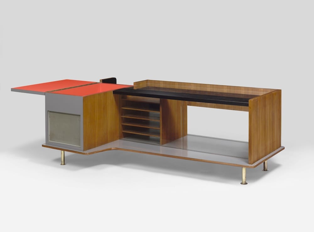 Mid-century modern wooden desk with red accents and open shelves on a gray background.