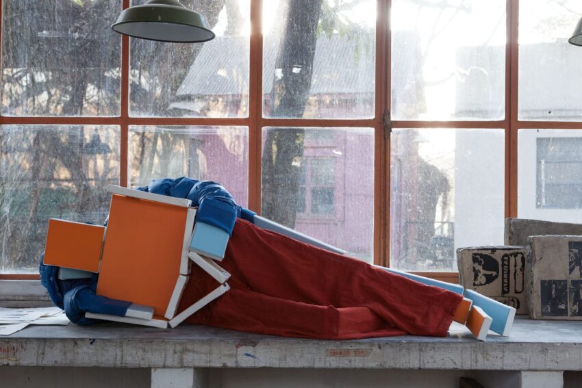 Sculpture of a geometric humanoid figure with red pants and blue top lying on a tabletop in a sunlit room.