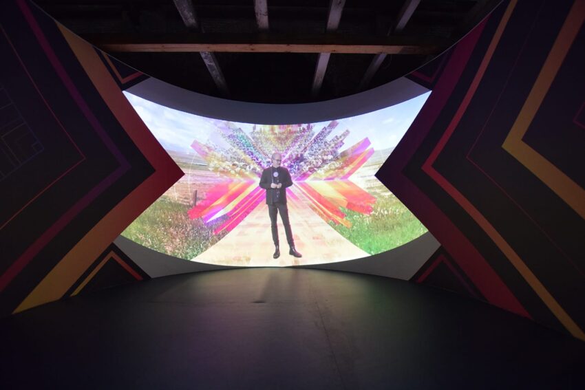 Person standing in front of a colorful, abstract digital projection on a curved screen inside a dimly lit room.
