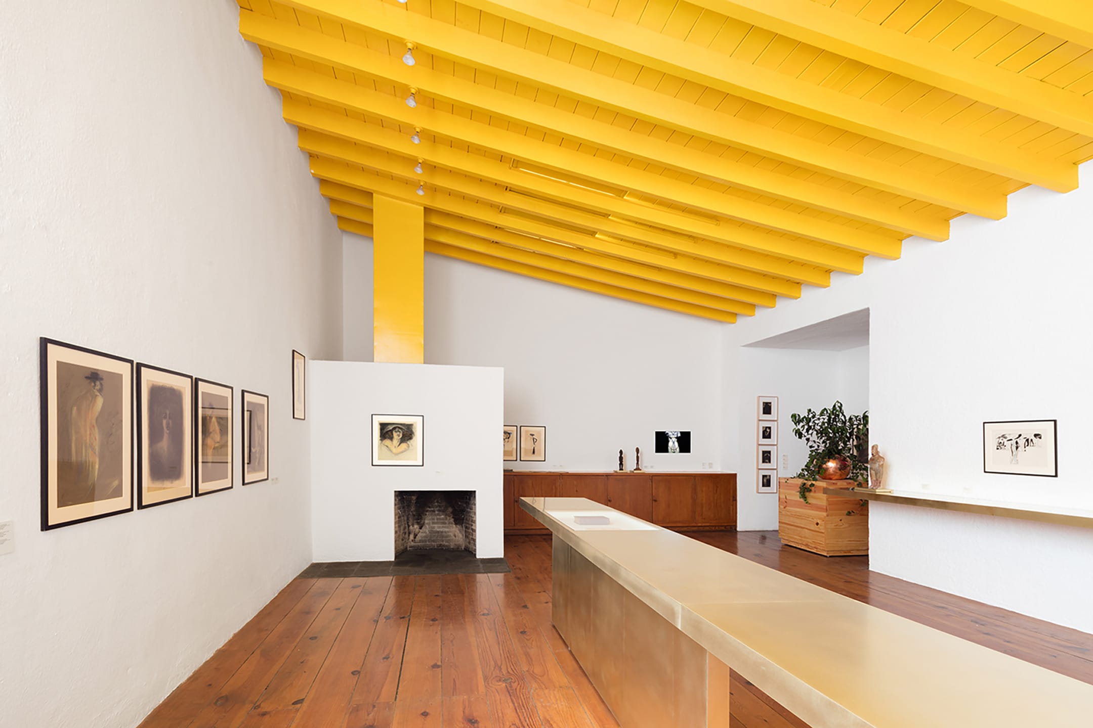 Modern art gallery with yellow ceiling, wooden floor, framed artwork on white walls, and a central minimalist table.