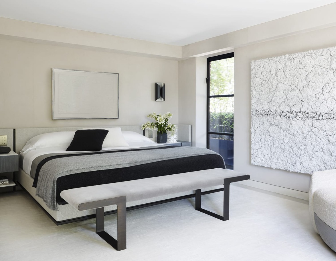 Modern bedroom with a large bed, gray tones, a bench at the foot, abstract art on the walls, and a window view.