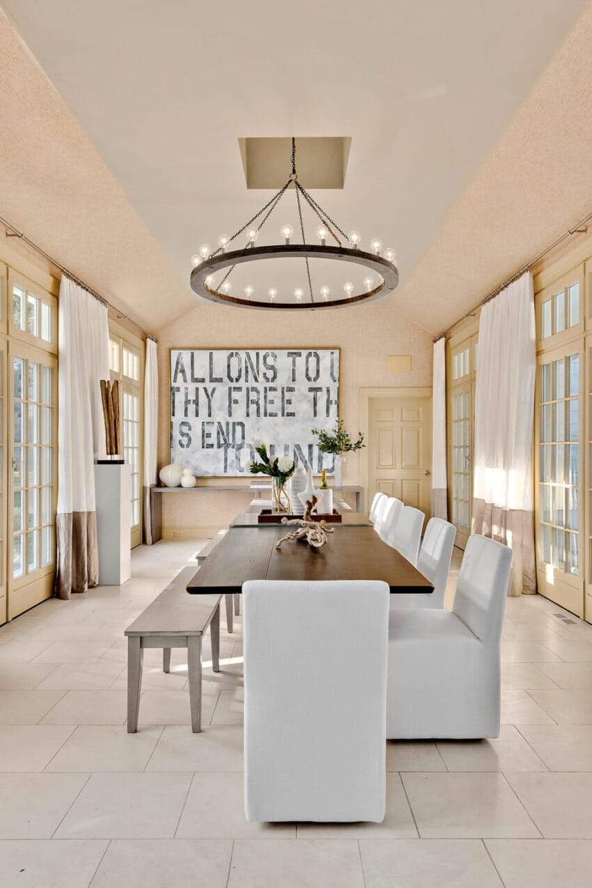Elegant dining room with large chandelier, wooden table, white chairs, and modern artwork on creme walls with large windows.