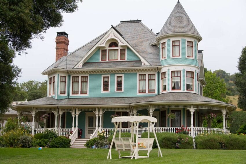 Victorian-style house with a turret and wraparound porch, surrounded by green lawn and trees, with a white garden swing.