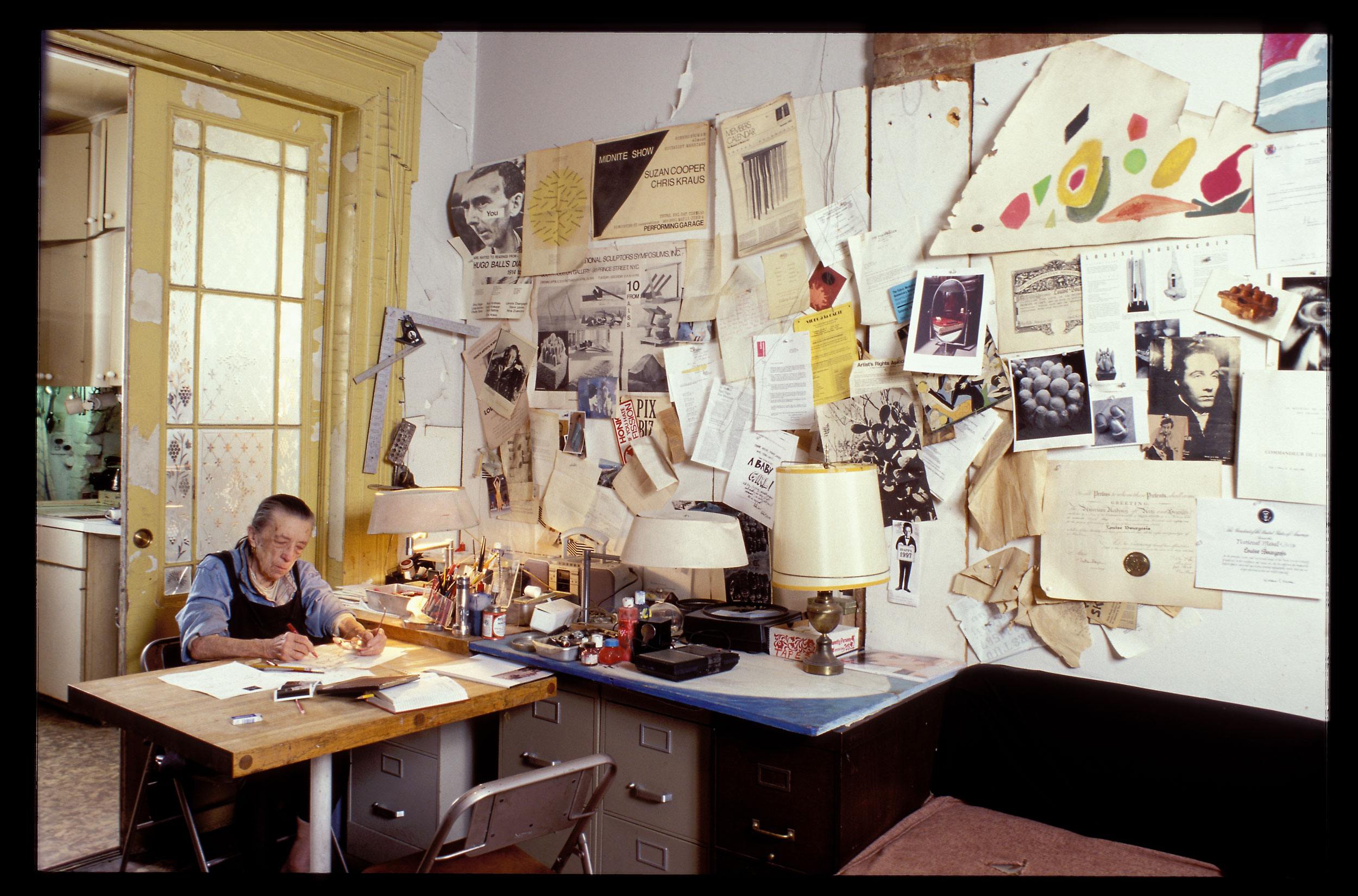 Artist working at a cluttered desk surrounded by art and notes on the wall in a studio space.