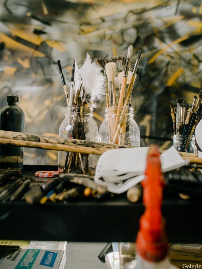Art supplies on a cluttered table with paintbrushes in jars, a paint roller, and various painting tools in a creative studio.