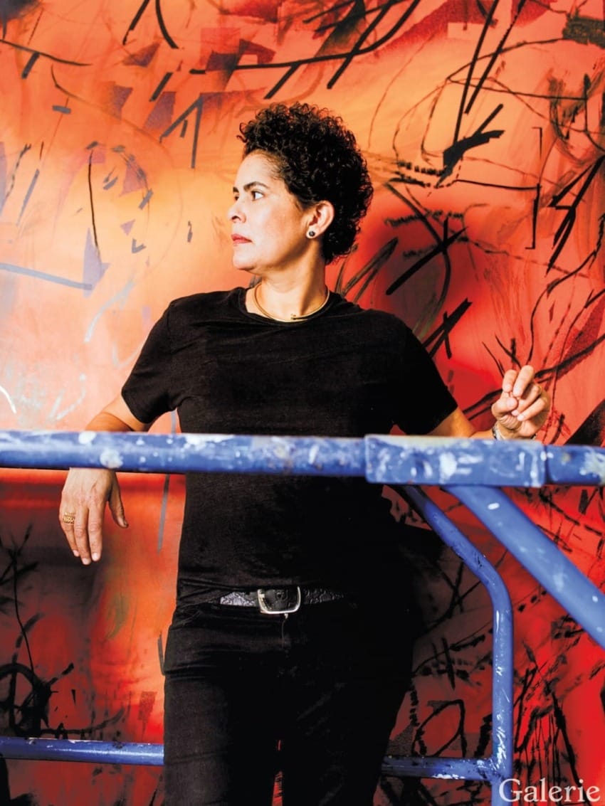 Artist standing in front of a vibrant abstract mural, wearing a black shirt, with a thoughtful expression.