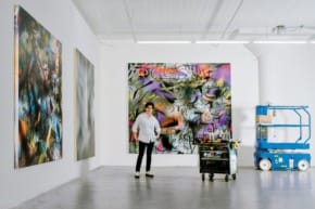 Artist standing in a studio with vibrant abstract paintings on the walls, beside a mobile workstation and a blue lift.