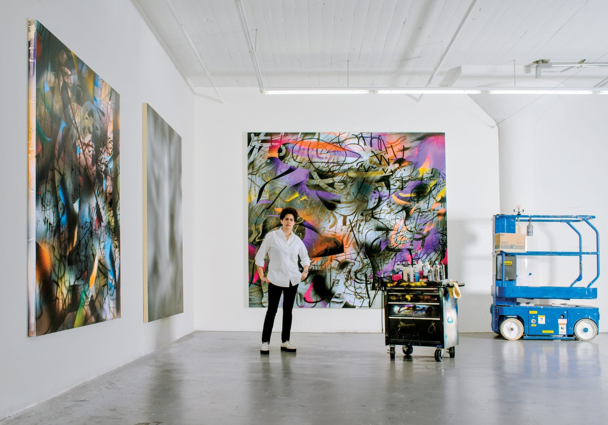 Artist standing in a studio with vibrant abstract paintings on the walls, beside a mobile workstation and a blue lift.