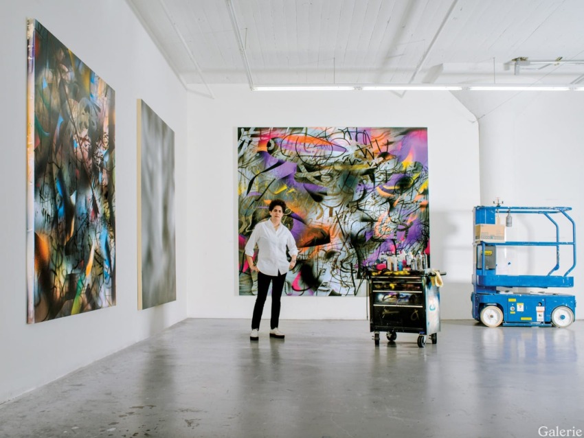 Artist standing in a studio with vibrant abstract paintings, next to a cart with art supplies and a blue lift in the background.