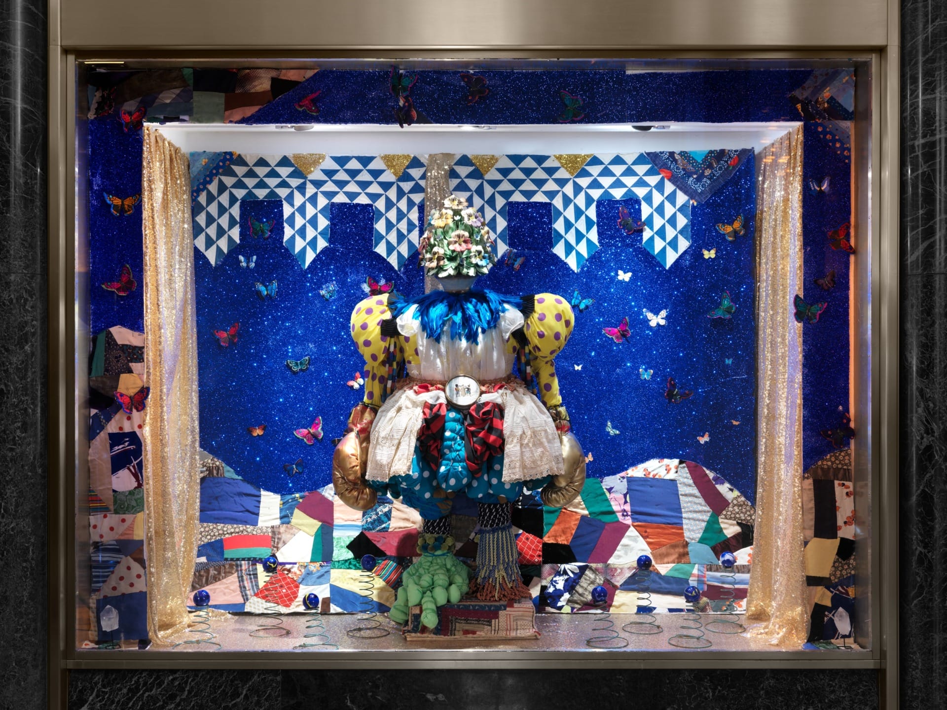 Colorful, whimsical window display with bold patterns, vibrant fabrics, and decorative elements against a blue backdrop.