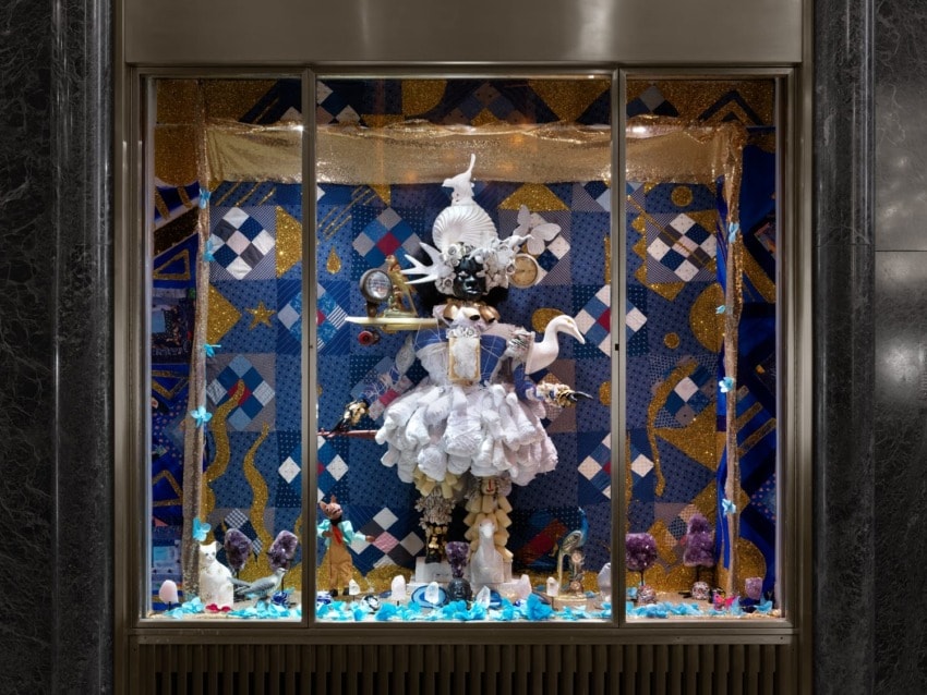 Elaborate store window display with a mannequin in a white dress, geometric patterns, and various decorative items.