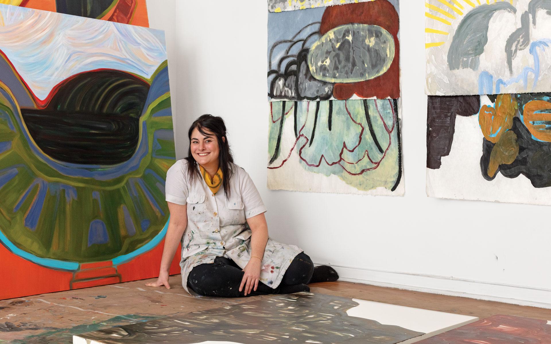 Artist sitting on the floor surrounded by colorful abstract paintings in a studio setting.