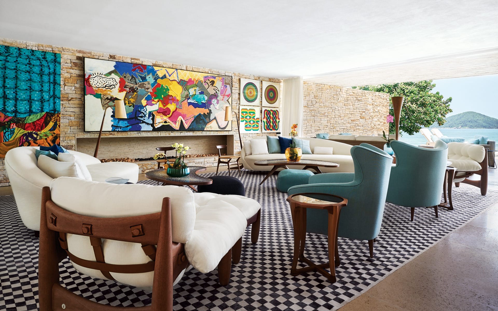 Spacious living room with modern art, white sofas, blue armchairs, wooden coffee tables, and a scenic view through large windows.