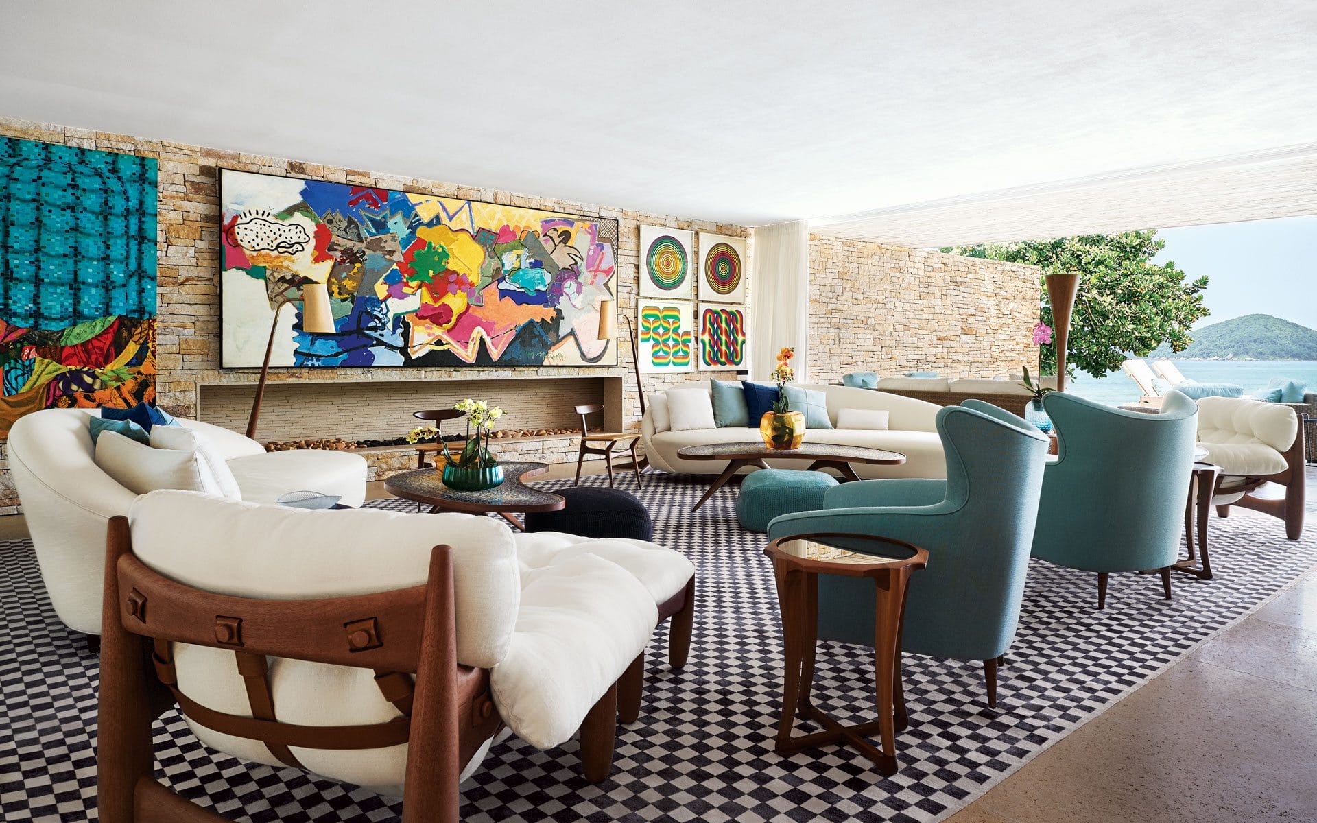 Modern living room with abstract art, white and teal furniture, checkered floor, and ocean view through large windows.