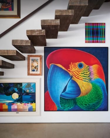 Colorful paintings, including a large parrot artwork, displayed on a white wall under wooden stairs.