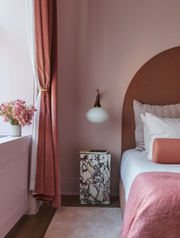 Cozy pink bedroom with velvet curtains, marble side table, bed with plush pillows, and a modern pendant light.