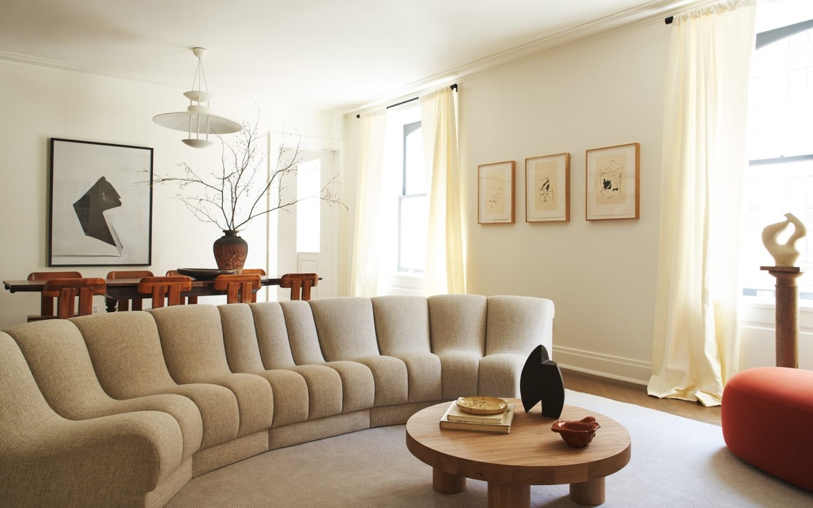 Modern living room with curved beige sofa, wooden table, abstract art, and large windows with sheer curtains.
