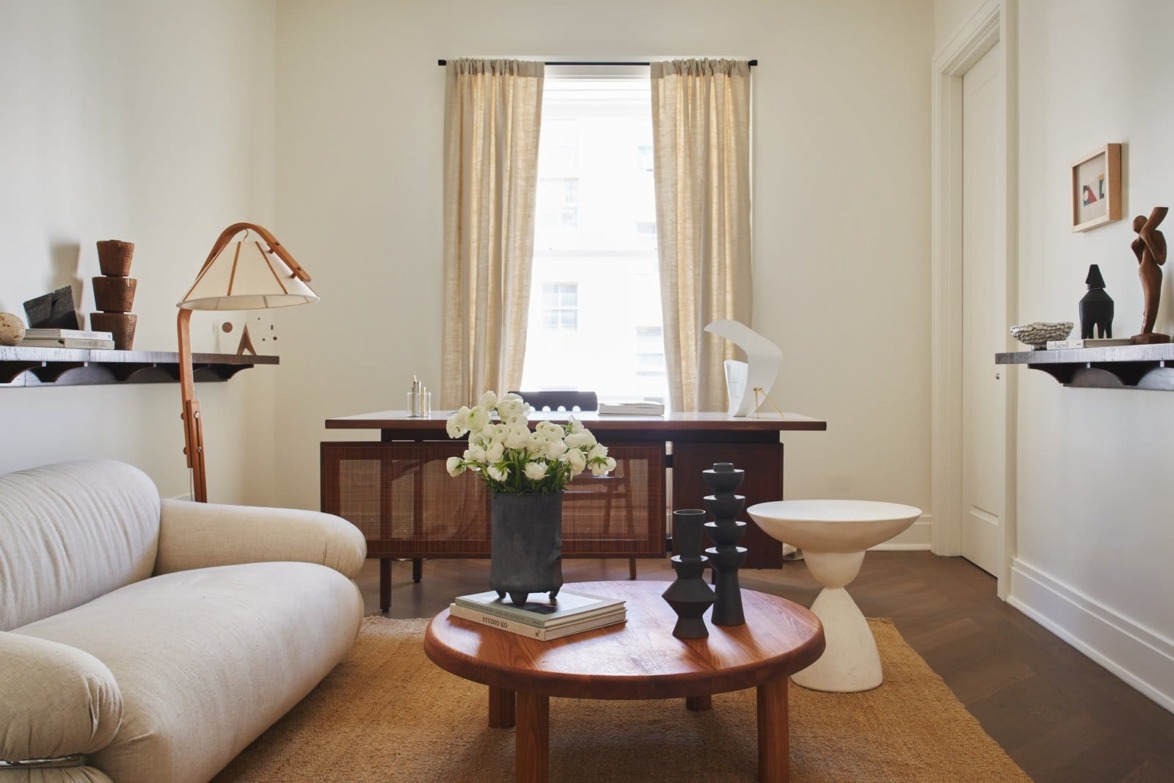 Minimalist home office with a beige sofa, wooden desk, and artistic decor, featuring a round coffee table with flowers.