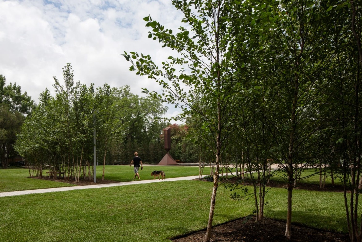 Person walking a dog on a path through a lush green park with tall trees and cloudy skies in the background