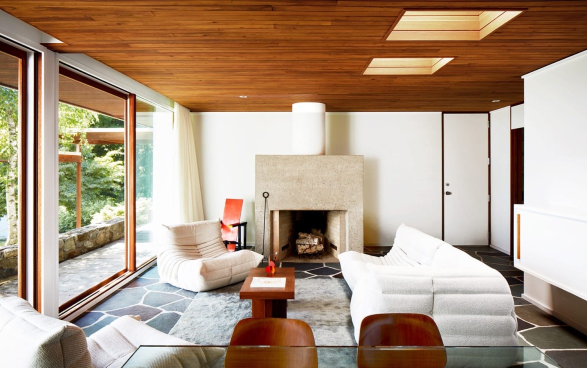 Modern living room with large windows, wooden ceiling, stone fireplace, white sofas, and glass table overlooking greenery.
