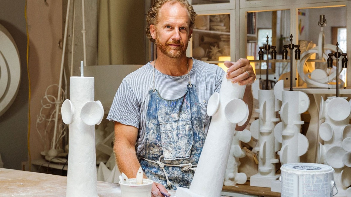 Artist working with white sculptural pieces in a studio filled with various art materials and tools.