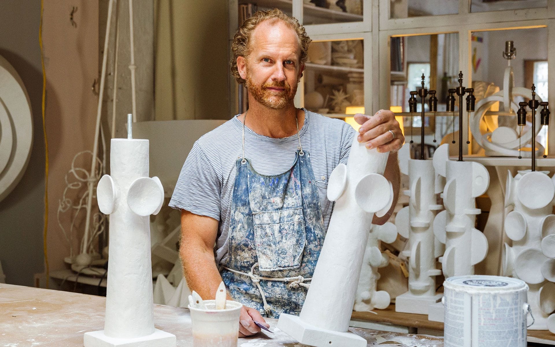 Artist working with white sculptural pieces in a studio filled with various art materials and tools.