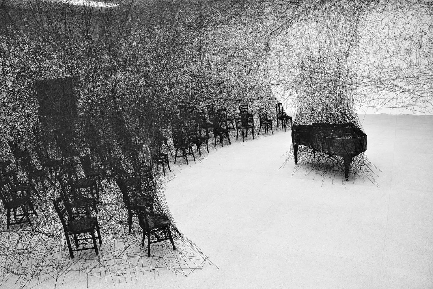Room filled with black threads, numerous chairs, and a grand piano, creating an intricate and artistic installation.