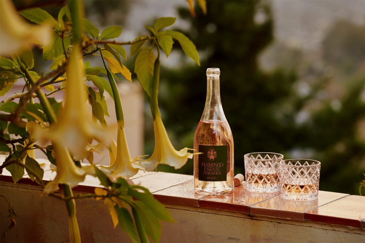 Bottle of Flamingo Estate wine and two glasses on an outdoor ledge with yellow flowers in the foreground.