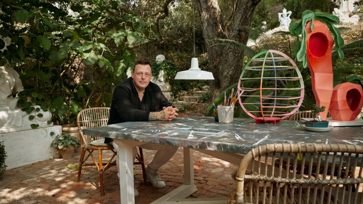 Man sitting at an outdoor table surrounded by artistic decorations and plants in a garden setting.