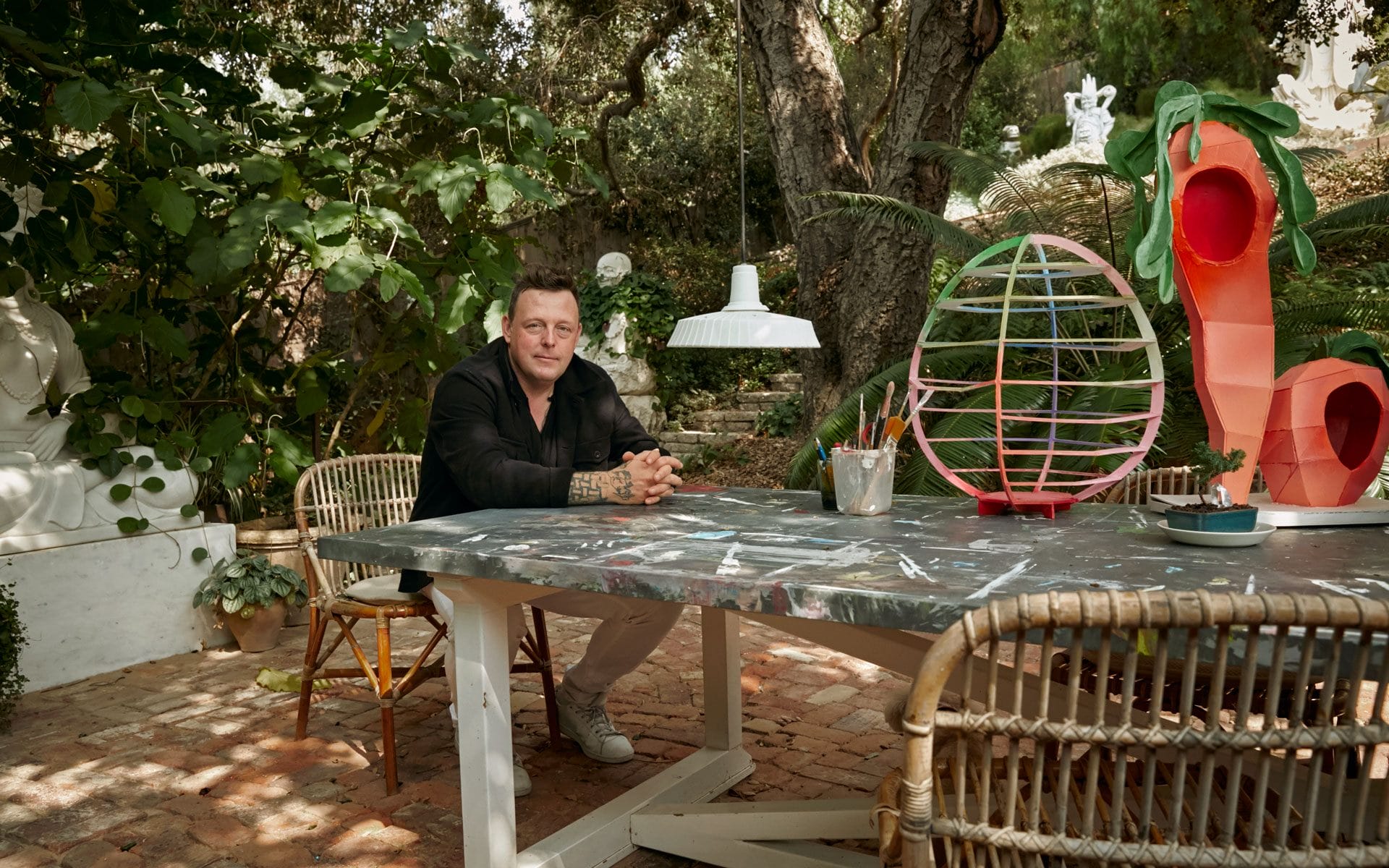 Man sitting at an outdoor table surrounded by artistic decorations and plants in a garden setting.