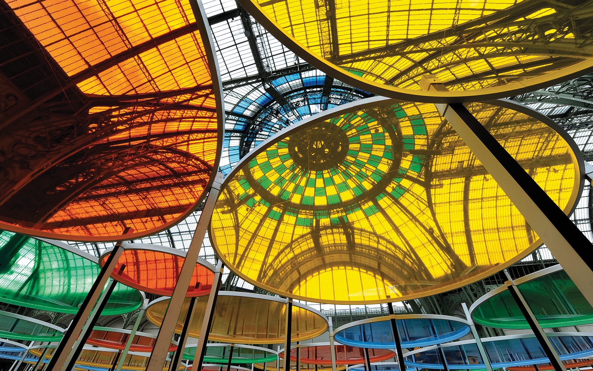 Colorful circular canopies with stained glass effect inside a large architectural structure with a glass roof.