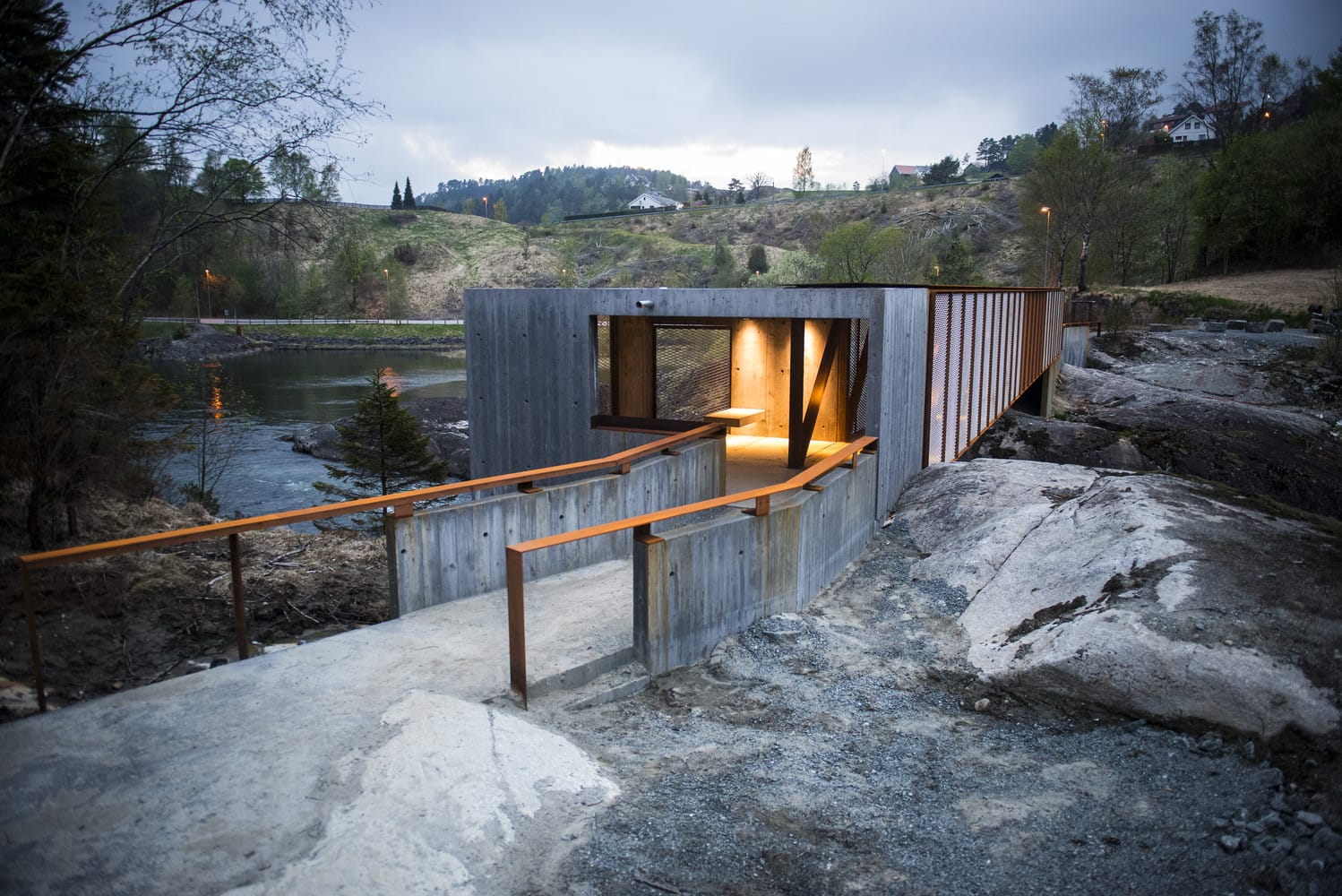 Modern wooden and concrete building with a pathway, surrounded by a natural landscape and river.