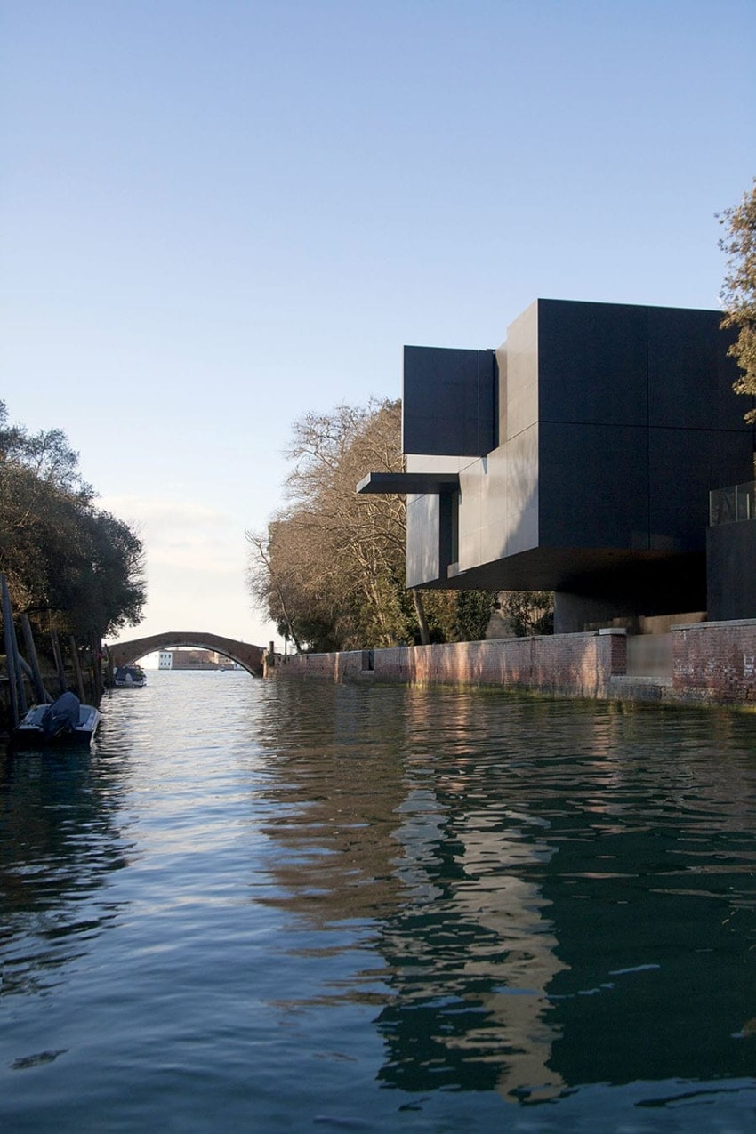 Modern black building along a canal with trees lining the waterway and a distant bridge under a clear blue sky.