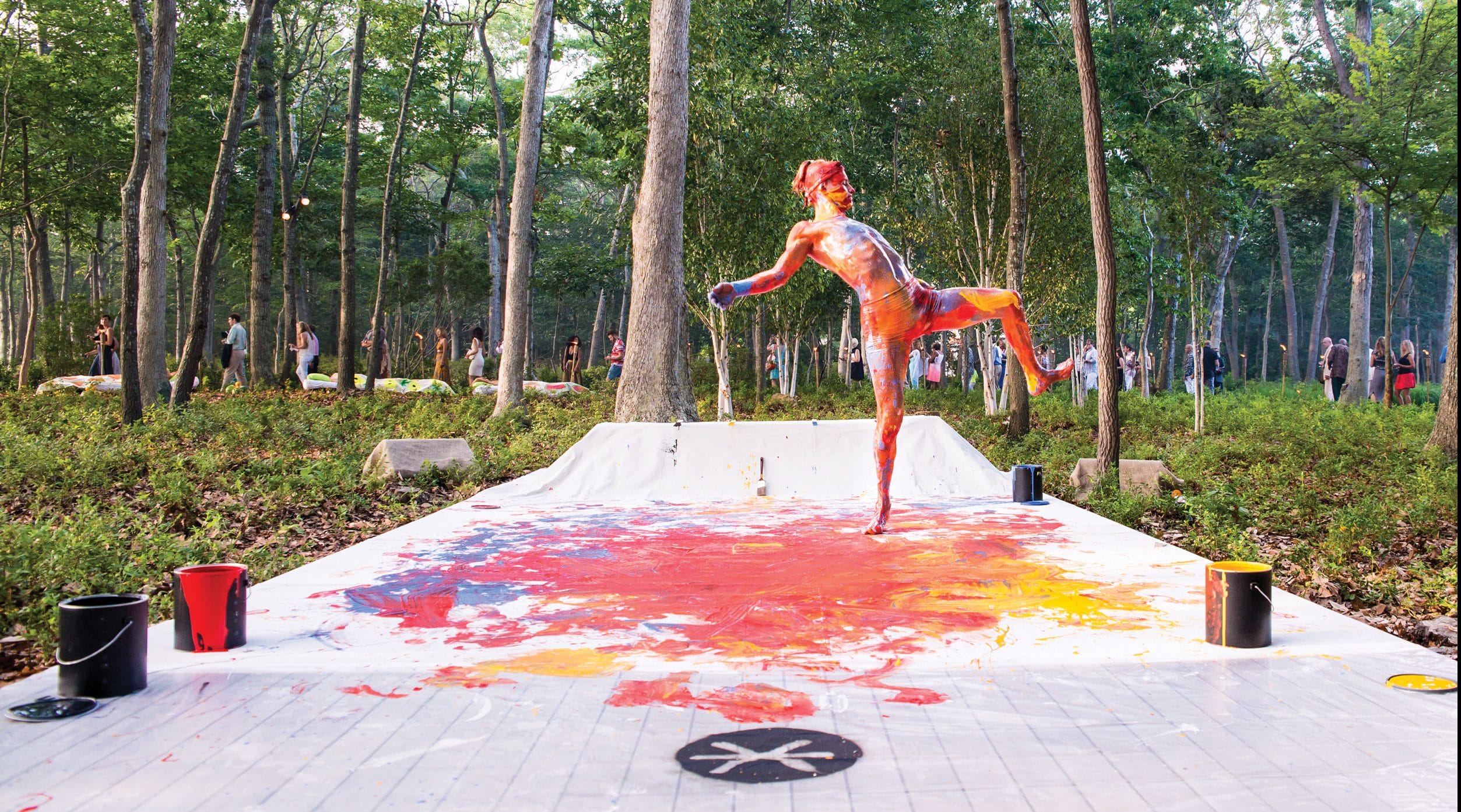 Person covered in colorful paint dancing on paint-splattered floor in forest setting with onlookers in the background.