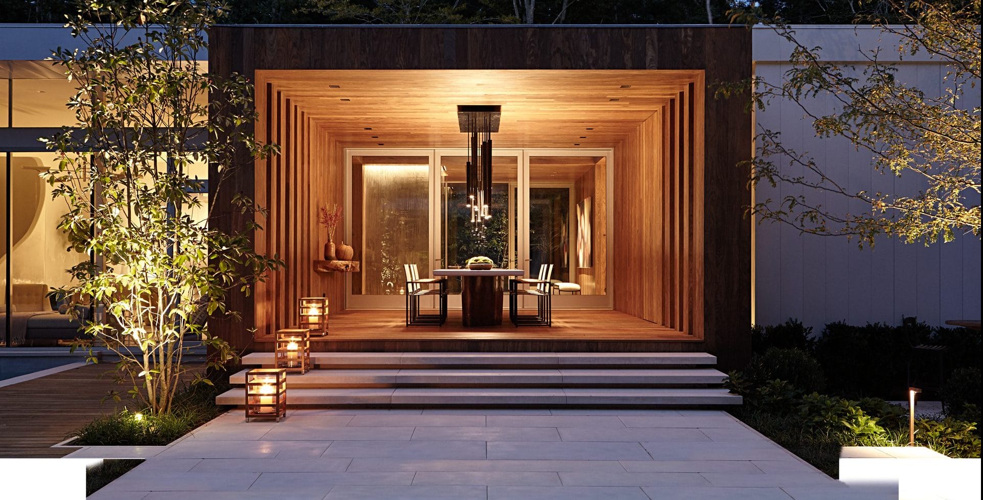 Modern outdoor dining area with wood accents, glass walls, and lanterns, surrounded by trees and soft lighting in the evening.